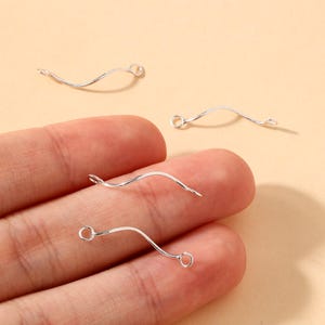May include: Four silver-colored, wavy earring hooks. Each hook has a small loop on one end and a pointed tip on the other. The hooks are displayed against a neutral background, with some held in a person's hand.