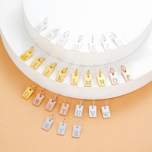 May include: A collection of rectangular initial charms in gold, silver, and rose gold. Each charm features a single letter from the alphabet, with a small loop for attachment. The charms are displayed on a white, tiered surface.