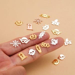 Op de afbeelding: Een verzameling kleine metalen bedels in goud- en zilverkleur met Halloween-thema's zoals schedels, spinnenwebben, geesten, pompoenen, grafstenen, katten en het woord BOO, voorzien van kleine oogjes voor bevestiging aan sieraden.