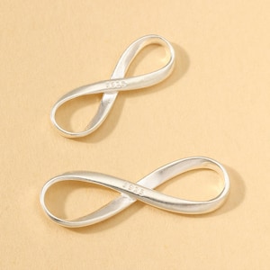 May include: Two silver-colored infinity charms. Each charm is shaped like a figure eight, with a smooth, polished surface. The charms are marked with "925" on the surface. The background is a solid, light tan color.