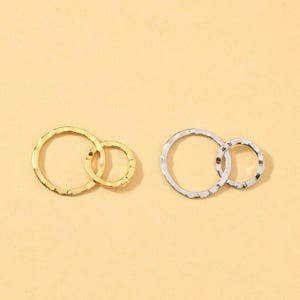 Puede incluir: Dos pares de pendientes de círculo enlazados. Un par es dorado, el otro es plateado. Cada pendiente presenta dos círculos entrelazados con una textura martillada. Los pendientes se muestran sobre un fondo beige claro.
