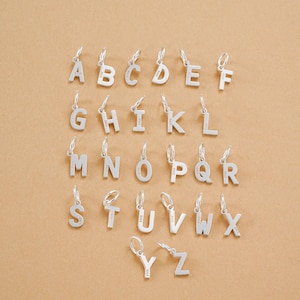 May include: Silver alphabet charms, each with a small loop for attaching to a necklace or bracelet. The letters A through Z are displayed, showcasing a variety of fonts and styles. The charms are arranged on a neutral, light brown surface.