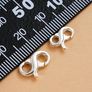 May include: Two silver-colored, figure-eight shaped jewelry clasps. The clasps are positioned on a tan surface next to a black ruler with white markings. The clasps have a small hook on one end and a loop on the other, designed for securing jewelry.
