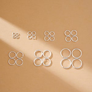 May include: A collection of silver-colored hoop earrings in various sizes, arranged on a light brown surface. The earrings are circular and appear to be made of metal. The image showcases the earrings in sets of two, creating a symmetrical display.