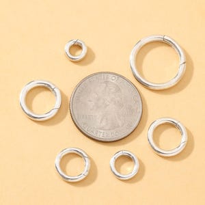 May include: A collection of silver-colored, round clasps in various sizes, arranged around a quarter coin. The clasps have a simple, circular design with a hinged opening. The background is a solid, light tan color.
