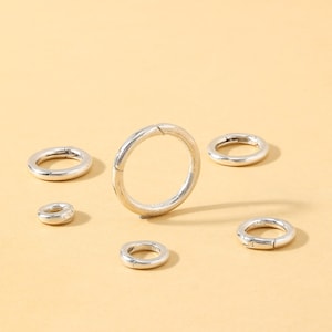 May include: A collection of silver-colored, circular clasps of varying sizes, arranged on a light yellow surface. The clasps have a simple, minimalist design, with a hinged opening. The image is well-lit, highlighting the metallic sheen of the clasps.