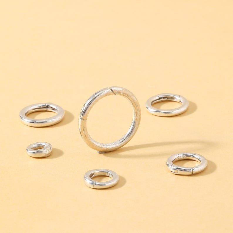 May include: A collection of silver-colored, circular clasps of varying sizes, arranged on a light yellow surface. The clasps have a simple, minimalist design, with a hinged opening. The image is well-lit, highlighting the metallic sheen of the clasps.