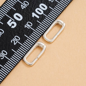 Puede incluir: Dos cierres rectangulares plateados están sobre una superficie marrón claro, junto a una regla negra con marcas blancas. Los cierres están abiertos, mostrando su estructura interna. La regla muestra medidas en centímetros.