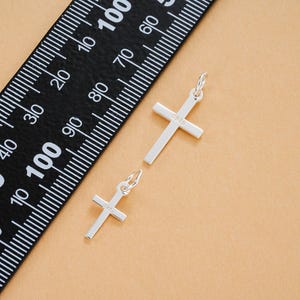 May include: Two silver cross pendants with a small loop at the top for a chain. The crosses are lying on a tan surface next to a black ruler with white markings. The crosses have the text "S925" on them.