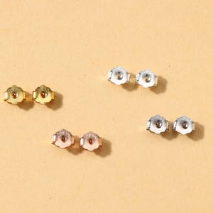 May include: A selection of earring backs in gold, silver, and rose gold hues. The backs are flower-shaped, with a central post hole. The earring backs are displayed against a light tan backdrop.