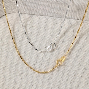 May include: Two delicate necklaces, one gold-toned and one silver-toned, are shown against a neutral backdrop. Each necklace has a textured, twisted chain design and a secure clasp. The gold necklace is positioned below the silver one.
