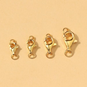 May include: Four gold-colored lobster claw clasps of varying sizes. Each clasp has a spring-loaded mechanism and a small ring for attaching to jewelry or other items. The clasps are arranged on a light tan background.