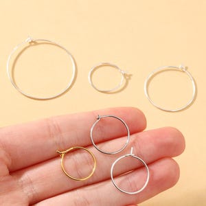 Puede incluir: Una colección de pendientes de aro en varios tamaños y colores. Los pendientes son plateados y dorados, con un diseño sencillo y minimalista. Los aros se muestran sobre un fondo amarillo claro.