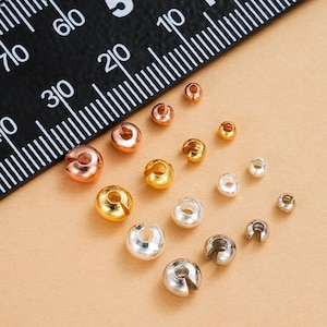 May include: An assortment of crimp beads in various metallic finishes, including gold, silver, and copper. The beads are of different sizes and shapes, with a ruler in the background. These jewelry-making components are used to secure clasps and other findings.