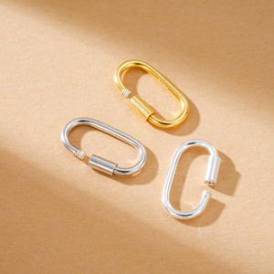 Op de afbeelding: Drie ovale karabijnsluitingen in goud en zilver. De gouden sluiting bevindt zich rechtsboven, de zilveren eronder. Ze hebben een schroefvergrendelingsmechanisme. De achtergrond is effen beige.