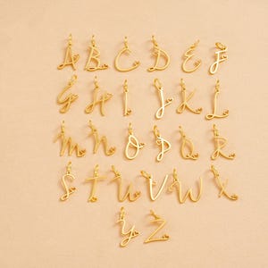 Op de afbeelding: Een verzameling goudkleurige alfabetbedels, elk in een cursief lettertype. Elke letter heeft een klein hartdetail. De bedels worden gepresenteerd op een licht perzikkleurig oppervlak.
