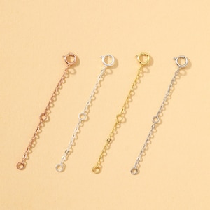 Puede incluir: Cuatro extensores de cadena metálicos en tonos oro rosa, plata y oro. Cada extensor presenta una pequeña cadena con un cierre en un extremo y un anillo en el otro, diseñado para alargar collares o pulseras. El fondo es de color beige claro liso.