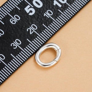 Może przedstawiać: Srebrny, owalny zatrzask do biżuterii. Zatrzask znajduje się obok czarnej linijki z białymi oznaczeniami. Zatrzask ma około 2 cm długości.