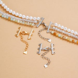 Könnte beinhalten: Nahaufnahme von Schmuckverschlüssen und Halsketten. Die Verschlüsse sind gold- und silberfarben, mit kleinen, klaren Steinen. Die Halsketten zeigen weiße Perlen und bunte Perlen. Der Hintergrund ist einfarbig beige.