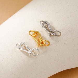 Puede incluir: Tres cierres de gancho metálicos en tonos plateado y dorado. Los cierres tienen un diseño decorativo con un gancho curvo y un extremo en bucle. El cierre dorado tiene una superficie texturizada. Los cierres son probablemente para la fabricación de joyas o manualidades.