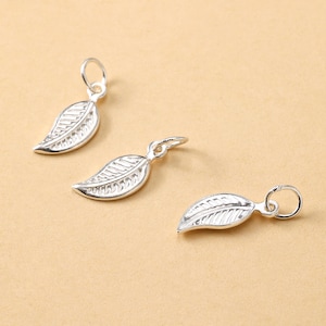 May include: Three silver leaf charms with detailed veining and small loops for attaching to jewelry. The charms are displayed on a light brown surface, showcasing their reflective quality and delicate design.