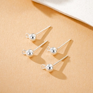 Puede incluir: Cuatro pendientes de plata con un pequeño diseño de bola redonda. Cada pendiente tiene un poste para la inserción y un pequeño lazo unido a la bola. Los pendientes se muestran sobre una superficie marrón claro.