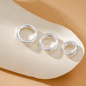 Pode incluir: Três fechos redondos prateados de tamanhos variados são exibidos em uma superfície de tom neutro. Os fechos têm um acabamento liso e polido e um design articulado para fácil abertura e fechamento. Os tamanhos variam de grande a pequeno.