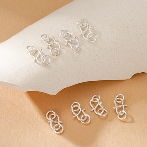 Puede incluir: Una colección de cierres de joyería plateados, dispuestos sobre una superficie de tono neutro. Los cierres presentan un diseño de gancho y ojo, ideales para collares y pulseras. Los cierres están hechos de un material metálico brillante.