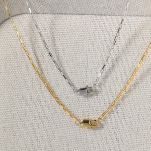 Pode incluir: Duas delicadas correntes com elos de corrente tipo caixa. Uma é dourada, a outra prateada. Ambas as correntes têm fecho de lagosta. As correntes são exibidas em uma superfície texturizada esbranquiçada.