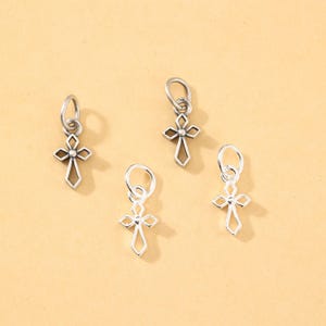 Può includere: Quattro piccoli ciondoli a forma di croce color argento con disegni traforati sono esposti su uno sfondo beige chiaro. Ogni ciondolo ha un anello nella parte superiore per il fissaggio a una collana o un braccialetto. Le croci hanno un tema religioso classico.