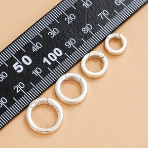 Könnte beinhalten: Vier silberfarbene, runde, klappbare Verschlüsse in verschiedenen Größen werden auf einer beigefarbenen Oberfläche gezeigt. Ein schwarzes Lineal mit weißen Markierungen liegt diagonal, als Größenreferenz. Jeder Verschluss ist mit "925" markiert.