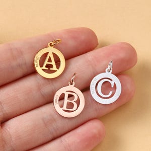 Könnte beinhalten: Drei gold- und silberfarbene Initialanhänger. Die Anhänger sind kreisförmig mit den Buchstaben A, B und C in der Mitte ausgeschnitten. Die Anhänger werden in einer Hand gehalten.