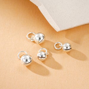 Op de afbeelding: Vier kleine, glanzende, zilverkleurige belletjes met een lusje aan de bovenkant. Elke bel heeft een gleuf in het midden. De bedels zijn gerangschikt op een lichtbruin oppervlak met een wit, getextureerd element op de achtergrond.