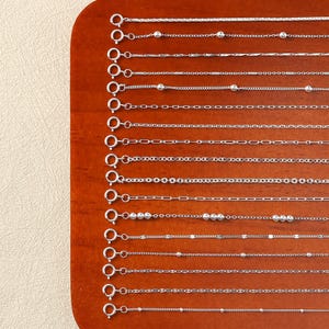 Può includere: Una collezione di collane d'argento esposte su una superficie di legno. Le collane presentano vari stili di catena, tra cui catene a sfere, a scatola e a cavo. Ogni collana ha una chiusura.