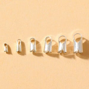 Peut inclure: Une collection de terminaisons de sertissage argentées de différentes tailles, disposées en rangée. Chaque élément a une boucle en haut et une base fendue et pliée. Les éléments sont présentés sur un fond beige clair.