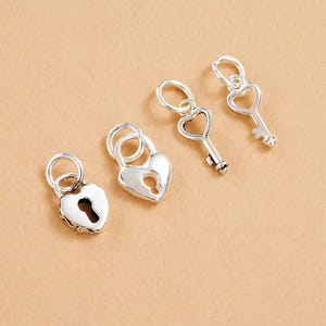 May include: Four silver charms: two heart-shaped padlocks and two keys. The padlocks have keyholes, and the keys have heart-shaped cutouts. All charms have a small loop for attaching to a bracelet or necklace. The background is a solid tan color.