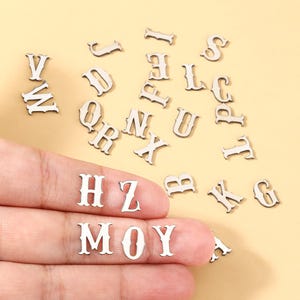 May include: A collection of small, silver-colored wooden alphabet letters in a serif font. Some letters are scattered on a light yellow surface, while others are held in a hand. The letters are used for crafting and personalization projects.