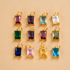 以下が含まれることがあります： さまざまな色の宝石がセットされた、12個の金色の長方形ペンダントのコレクション。宝石には、赤、紫、青、白、緑、ピンク、黄色が含まれます。ペンダントは、薄茶色の表面に配置されています。