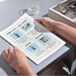 May include: A person holding a white printed sheet with illustrated instructions for balance and coordination exercises. The sheet features colorful drawings and text, including "Wobble Catch," "Roll & Collect," "Balance," and "Ball Passes."