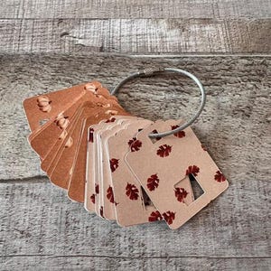 May include: A set of rectangular kraft paper tags held together by a silver metal cable ring. The tags feature a repeating pattern of small dark red autumn leaves and a square cutout at the bottom edge, with some tags showing pumpkin icons.