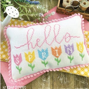 Puede incluir: Una almohada blanca con ribete rosa y un diseño de punto de cruz que dice "hello" con una fila de tulipanes coloridos debajo. La almohada está sobre una tela a cuadros amarilla y blanca. El texto "PRIMROSE COTTAGE STITCHES" está en la parte superior de la imagen y el texto "HELLO SPRING" está en la parte inferior de la imagen.