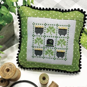 Op de afbeelding: Een groen en wit kussen met een zwarte bies heeft een kruissteekontwerp van een negen-patch patroon met klavertjesvier en potten goud. Het kussen heeft de titel "March Nine Patch" en is van "Lindsey Weight". De tekst "Primrose Cottage Stitches" staat onderaan de afbeelding.