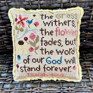 May include: A beige pillow with a yellow fringe border features a cross-stitch design with colorful flowers and the text "the grass withers, the flower fades, but the word of our God will stand forever. Isaiah 40:8".