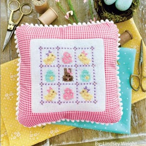 May include: A pink and white gingham pillow with a white embroidered square featuring a nine-patch design of chicks, eggs, and a bunny. The pillow is surrounded by white pom-poms and the text "April Nine Patch" is at the top of the image. The text "Primrose Cottage Stitches" is at the bottom of the image.