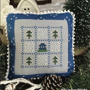 Puede incluir: Una almohada de punto de cruz azul y blanca con una escena de invierno que presenta una pequeña casa, árboles y nieve. La almohada tiene un borde de pompones blancos y está rodeada de ramas de pino. El texto "January Nine Patch" y "Primrose Cottage Stitches" es visible.