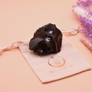 Könnte beinhalten: Ein dunkler, glänzender Obsidianstein liegt auf einem beigen Kordelzugbeutel mit goldfarbenem Text und einem Mond- und Augendesign. Der Hintergrund ist ein sanfter Pfirsichton, mit getrockneten lila Blumen rechts.