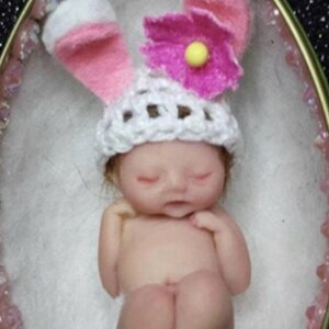 May include: A tiny, sleeping baby doll wearing a white crocheted hat with pink bunny ears and a pink flower on top. The doll is lying in a white egg-shaped container.