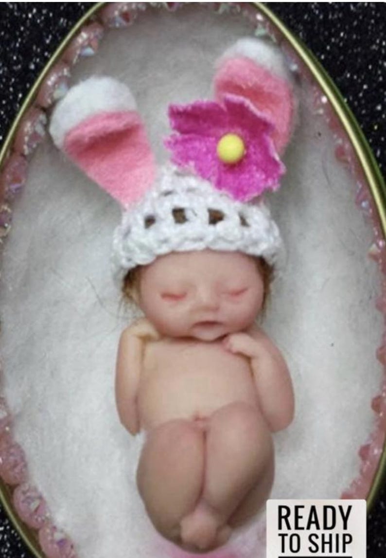 May include: A tiny, sleeping baby doll wearing a white crocheted hat with pink bunny ears and a pink flower on top. The doll is lying in a white egg-shaped container.