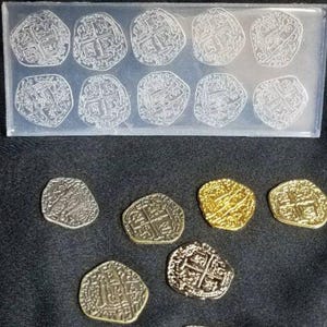 Op de afbeelding: Een verzameling replica munten, sommige in een doorzichtige mal, andere verspreid op een donkere ondergrond. De munten variëren in kleur, waaronder zilver- en goudtinten, en hebben ingewikkelde kruis- en embleemontwerpen.