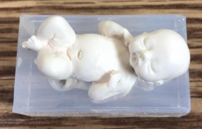 May include: A white resin sculpture of a baby lying on its back in a clear plastic box.